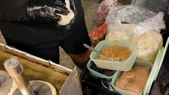 Close up shot of vendor hands in black gloves using a spoon to add brown sugar and nut filling into raw dough for traditional Hotteok sweet pancakes at a market.
