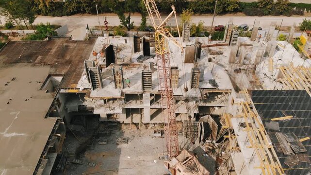 Aerial view of busy construction site with reinforced concrete slabs, exposed rebar, tower crane, timber formwork, support columns, workers, and unfinished building structure under warm daylight