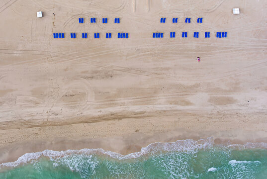 Aerial view of blue lounge chairs arranged on the sandy beach with tire tracks and turquoise ocean waves in Miami, Florida, United States.