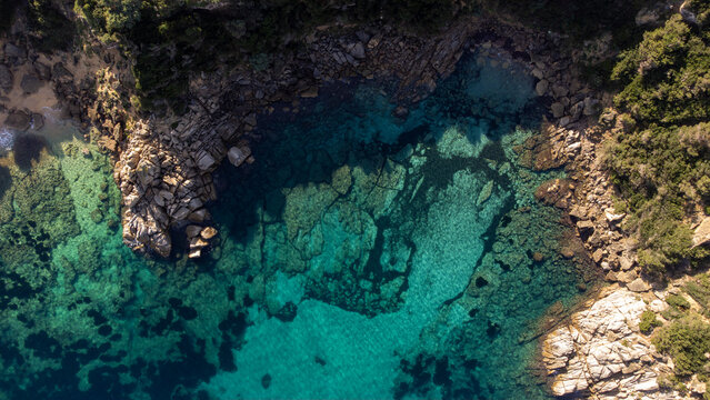 Aerial view of the rocky coastline and turquoise waters of Capo Testa, featuring clear seabed, rugged cliffs, and Mediterranean vegetation in Santa Teresa Gallura, Sardinia, Italy.