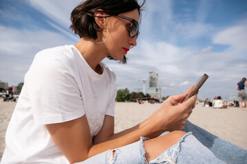 Side view of a millennial solo traveler using smartphone while sitting on a sandy beach, female digital nomad in sunglasses working on mobile phone near the sea during vacation © MarijaBazarova