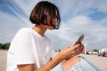 Side view of a millennial solo traveler using smartphone while sitting on a sandy beach, female digital nomad in sunglasses working on mobile phone near the sea during vacation © MarijaBazarova