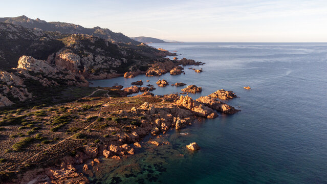 Aerial view of Cala Sarraina featuring rugged rocky cliffs, turquoise waters, and Mediterranean scrub along the coastline at golden hour in Trinita d'Agultu e Vignola, Sardinia, Italy.
