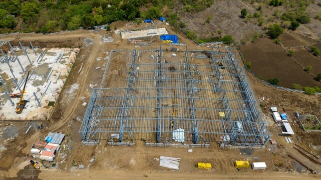 Aerial view of a warehouse construction site featuring a large steel frame structure, heavy machinery, and cranes on a dirt lot in Bhiwandi, Maharashtra, India.