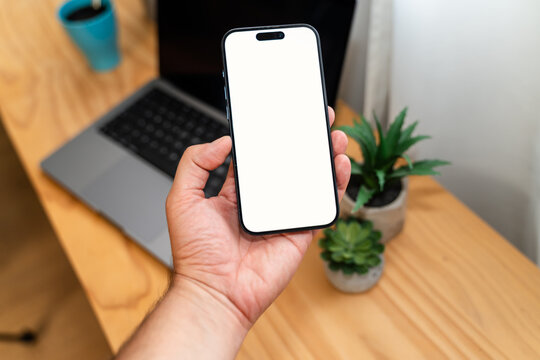 Hand holding smartphone with blank screen near laptop on desk