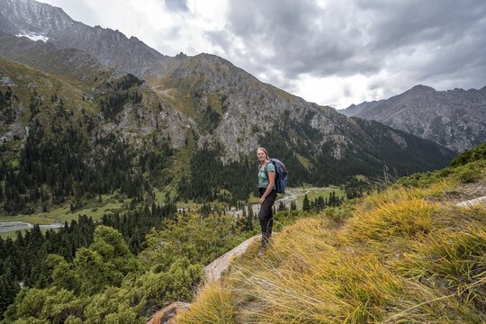 A female hiker stands on a grassy slope against an impressive mountain backdrop under a cloudy sky, Chong Kyzyl Suu Valley, Terskey Ala Too, Tien-Shan Mountains, Kyrgyzstan