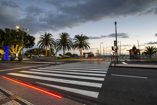 Evening scene at an illuminated pedestrian crossing with integrated LED floor marking, flanked by palm-lined streets and a brightly lit waterfront with Ferris wheel and pavilion. Funchal, Madeira, Portugal