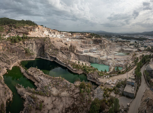 Aerial view of Cantera Porrino, a large granite quarry with deep green water pools, rocky cliffs, and industrial extraction areas under a cloudy sky, O Porrino, Pontevedra, Spain.