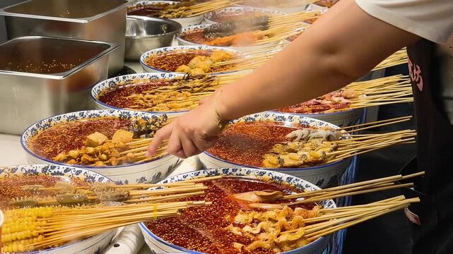 vendor hand choosing spicy skewer from large bowl of red oil broth. delicious street food with meat and seafood is popular in china. authentic street food attracts many hungry tourists