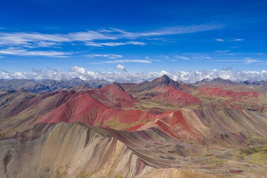 Aerial view of Vinicunca Rainbow Mountain with vibrant mineral-rich striped slopes, rugged Andean terrain, and distant peaks under a clear blue sky. Cusco, Cuzco, Peru.