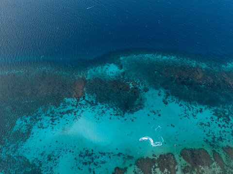Aerial view of a vibrant coral reef with turquoise shallow waters transitioning into deep blue sea, featuring a small boat wake in Karimunjawa, Central Java, Indonesia.