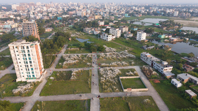 Aerial view of a residential area in dhaka, bangladesh, featuring unfinished apartment buildings, green spaces, and patches of white kash flowers