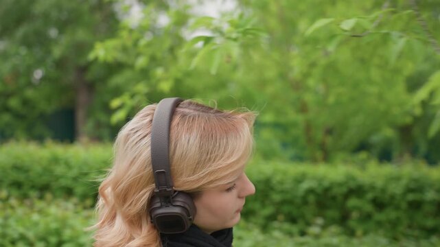 Lady walks through greenery while enjoying her headphones. Woman wears casual outerwear suitable for mild weather. background features lush foliage and blurred fence suggesting scene takes