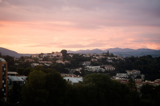 View of the hillside residential area at sunset, featuring houses nestled among trees with distant mountains under a soft pink sky in Nice, Provence-Alpes-Cote d'Azur, France.