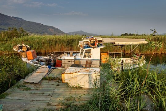 View of a small fishing boat docked at a wooden pier surrounded by tall reeds and mountains in the calm river of Akyaka, Mugla, Turkey.