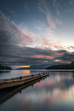 View of Lake Tarawera with a wooden jetty extending into the calm water under a dramatic sunset sky with clouds and distant mountains in Rotorua, Bay of Plenty Region, New Zealand.