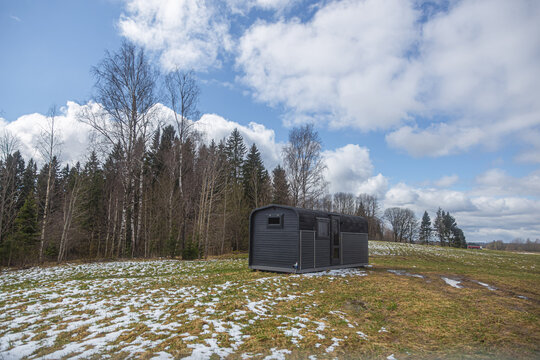 View of a modern dark-colored tiny house cabin in a rural field with patches of snow and a forest background in Latvia.
