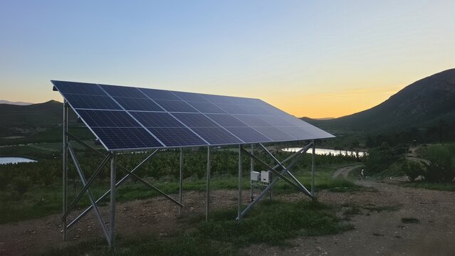 View of a solar panel array installed in a rural landscape with rolling hills, a small pond, and sunset lighting in Hoeko, Western Cape, South Africa.