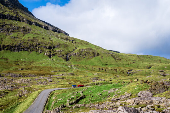 View of a red car with a trailer driving on a winding road through the mountainous green landscape of the Faroe Islands.