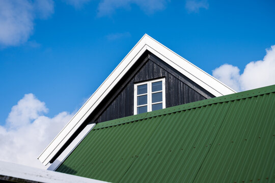 View of a house roof with green corrugated metal, black wooden gable, white trim, and a window against a blue sky with clouds in Faroe Islands.