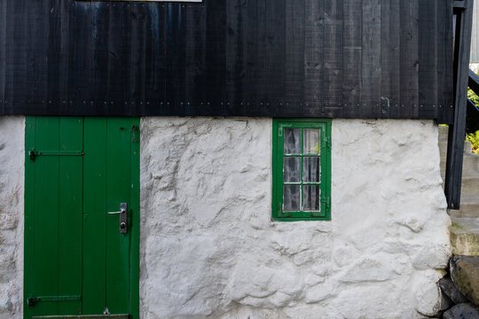 View of a traditional house with white stone walls, black wooden siding, a green door, and a green-framed window in the Faroe Islands.