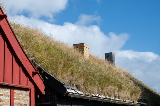 View of a traditional house with a grass-covered turf roof, featuring red and black wooden walls and chimneys under a blue sky in the Faroe Islands.