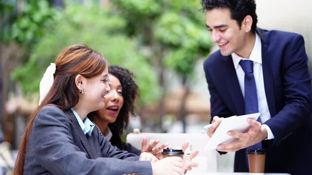 Three colleagues meet over a tablet at a shaded outdoor table, the man presenting while both businesswomen lean in, a compact alfresco project review in the city centre