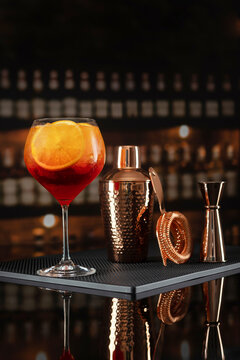 Orange cocktail with ice and citrus slice in glass. Copper shaker jigger strainer on black bar mat. Dark blurred bar background reflection