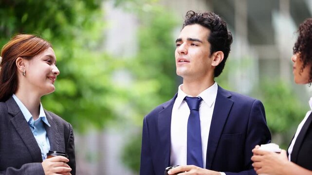 Close two-shot of a smiling businessman sharing a joke with two female colleagues over takeaway coffee, gesturing toward the skyline in a leafy business district