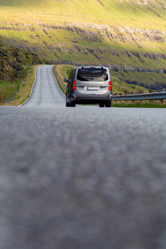 View of a silver van driving along a winding road through the green hills and rugged landscape of the Faroe Islands.