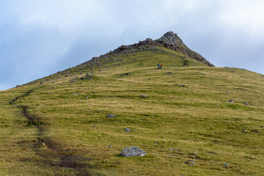 View of a grassy mountain slope with a hiking trail leading towards a rocky peak in the Faroe Islands.