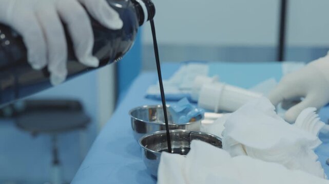 Surgical assistant pouring povidone-iodine antiseptic liquid into sterile metal bowl on operating table