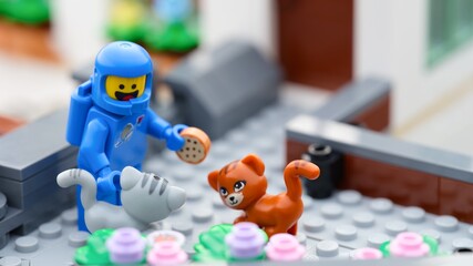 Obraz premium Tambov, Russian Federation - September 24, 2026 A blue Lego astronaut minifigure standing outside a house holding a miniature cookie, surrounded by a gray cat and a ginger cat.