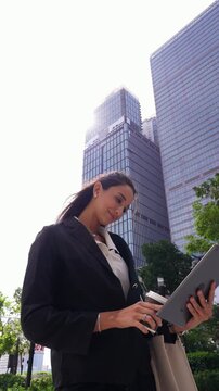 Vertical low angle shot of a Latina businesswoman reading a tablet beneath glass skyscrapers, sipping takeaway coffee and smiling against a bright sky in the city business district