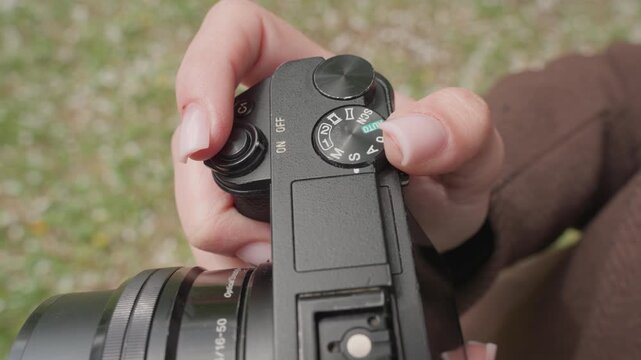 Close up view of hands adjusting camera settings focusing on dials and lens. Hand positioning indicates active engagement with device controls. Outdoor background suggests natural light