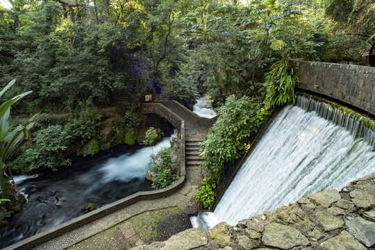 View of Parque Nacional Barranca del Cupatitzio featuring a stone bridge over a flowing river and a cascading waterfall surrounded by lush forest in Uruapan, Michoacan, Mexico.