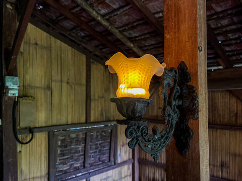 Vintage wall lamp with a fluted amber glass shade and ornate wrought iron bracket, glowing warmly against rustic wooden planks and a timber post in a cozy rustic interior.