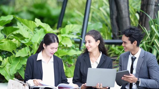 Three young business colleagues sit together in a green garden comparing a printed report with a laptop and a tablet, laughing and exchanging ideas as they review the numbers