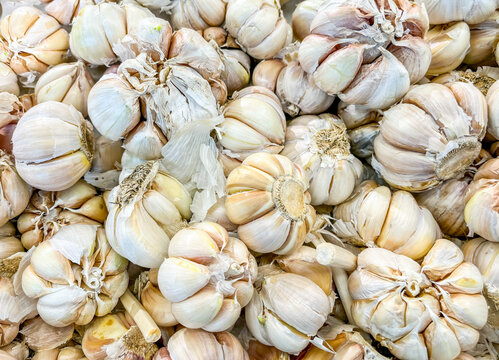 Fresh garlic bulbs pile closely together at a bustling traditional market stall, their papery white skins and dried brown stems densely filling the whole close-up frame.