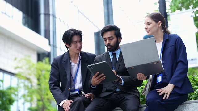 Smiling businesswoman shows a laptop screen to two male colleagues who hold a digital tablet, three professionals sitting on a stone ledge beside a glass office building on a sunny day