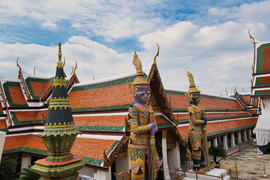 View of the ornate architecture and giant demon statues at The Grand Palace with traditional orange and green tiled roofs under a blue sky in Bangkok, Thailand.