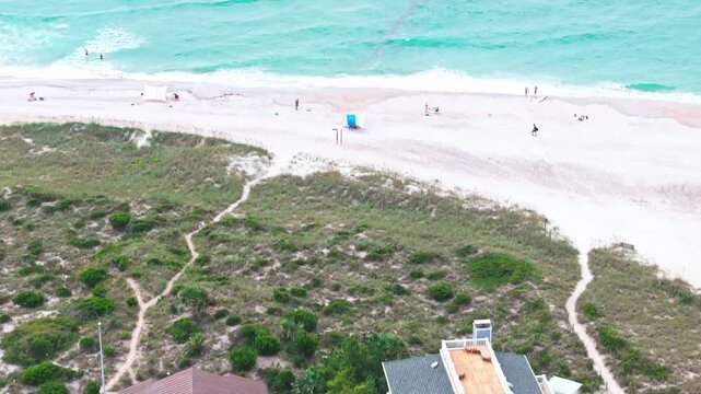 Aerial view of beachfront homes near turquoise ocean. Turquoise waves gently lap white sand. People stroll and sunbathe near shoreline. House features porches and colorful shutters