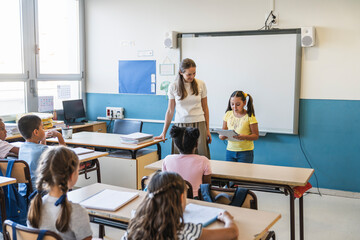 Young girl making a presentation to her classmates, supported by a female teacher in an elementary school classroom © Koldo_Studio