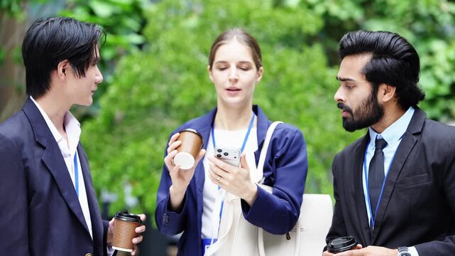 Businesswoman pulls her smartphone from a tote bag and shows the screen to two male colleagues, the bearded manager pointing at it as the team stands among green trees
