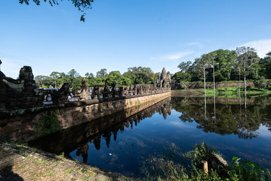 View of the historic stone causeway and moat leading to the Angkor Wat temple complex, surrounded by lush trees under a clear blue sky in Krong Siem Reap, Siem Reap Province, Cambodia.