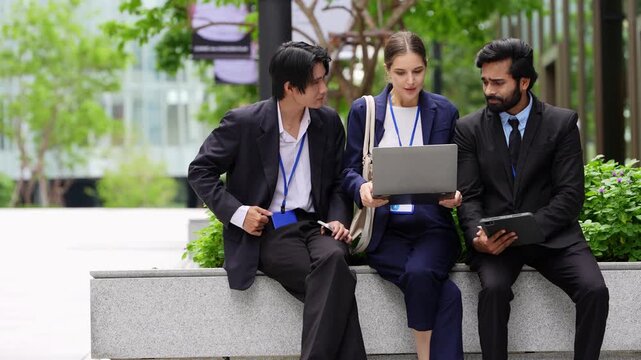 Wide view of three business colleagues sitting on a granite bench in a city plaza, a businesswoman holding an open laptop between two men who check a tablet and gesture