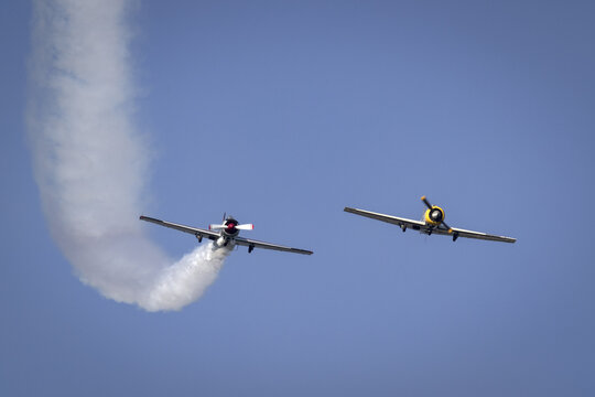 View of two vintage propeller aircraft performing an aerobatic maneuver with a smoke trail against a clear blue sky in La Spezia, Liguria, Italy.