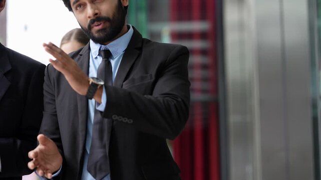 Three business colleagues walk past a glass shopfront talking, the bearded Indian man explaining with his hands, before the frame empties to show the window and city towers
