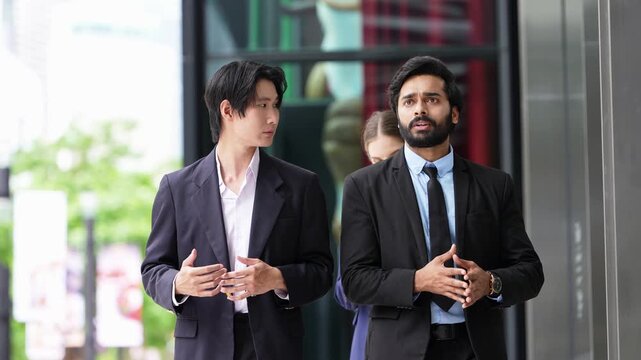 Asian businessman and bearded Indian colleague walk and talk past a shop window with a businesswoman carrying a tablet behind them, then turn toward each other to discuss