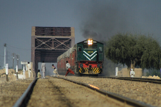 View of a freight train approaching a large steel bridge in the Cholistan Desert with people walking along the tracks in Bahawalpur, Punjab, Pakistan.